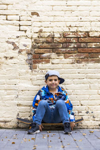 Portrait of smiling boy sitting on skateboard at footpath against wall