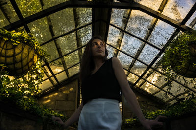 Low angle view of young woman looking up while standing in greenhouse