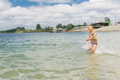 Side view of shirtless boy running in sea against cloudy sky during sunny day