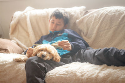 Boy with book sleeping while sitting by dog on sofa at home