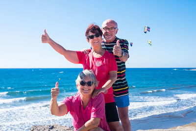 Portrait of smiling senior friends gesturing while at beach against sky