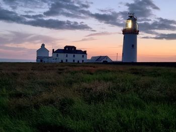 Lighthouse on field by building against sky during sunset