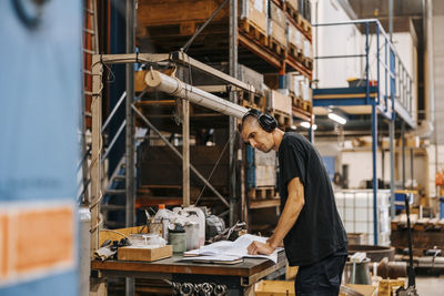 Male industry worker reading blueprint while wearing wireless headphones and standing near table in factory