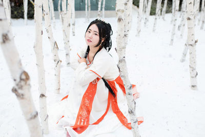 Side view portrait of young woman wearing traditional clothing while sitting by tree trunks during winter