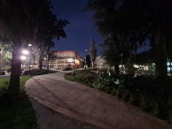 Illuminated street by buildings against sky at night