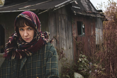 A grumpy woman in a warm head scarf and coat stands against the backdrop of an old abandoned house.