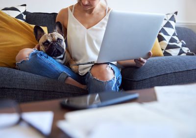 Midsection of woman with pug holding laptop while sitting on sofa at home