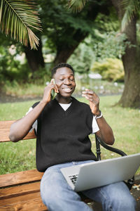 Young african man talking on phone, working on laptop in park, happy, pleased