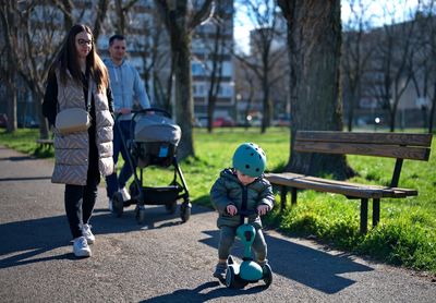 Little toddler on scooter and his parents with baby stroller walking in a park