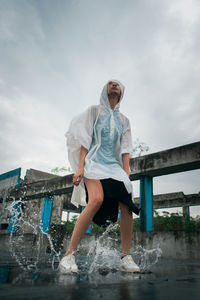 Full length of young woman wearing raincoat while jumping in puddle against sky