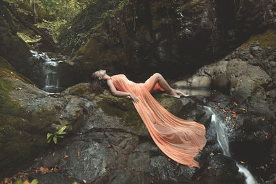 Teenage girl wearing orange dress while lying on rocks in forest