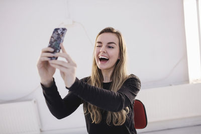 Cheerful female blogger winking while taking selfie through smart phone in office
