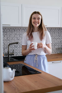 A cute girl of 15-18 years old stands in the kitchen with a mug in her hands