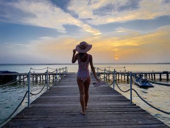 Woman wearing swimwear and summer hat walking down a wooden pontoon towards the sea at sunset