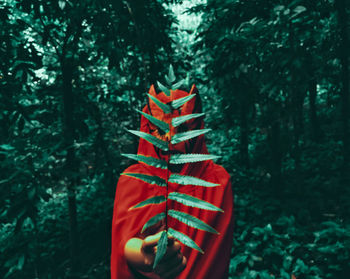 Person wearing red mask and cape holding plant against trees in forest