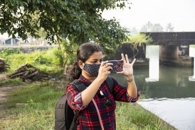 Woman wearing mask photographing river while standing against bridge