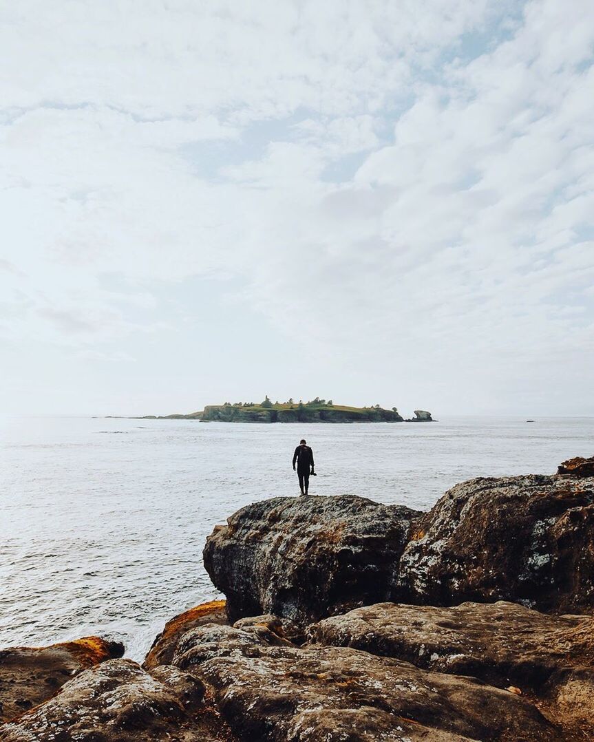 REAR VIEW OF MAN STANDING ON SHORE AGAINST SKY