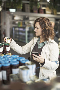 Woman holding smart phone while reading label on bottle in supermarket