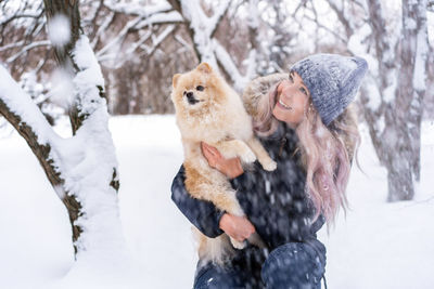 Young woman carrying dog while crouching in snow during winter