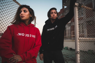 Couple wearing hoodie standing by fence against sky