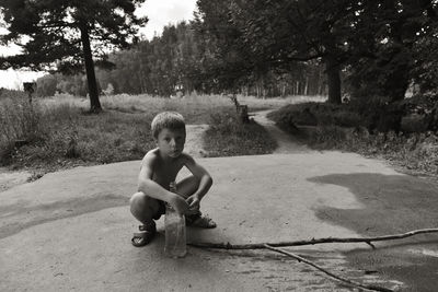 Portrait of shirtless boy with bottle crouching against trees