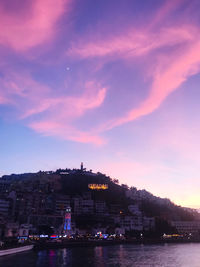 Illuminated buildings by sea against sky at sunset