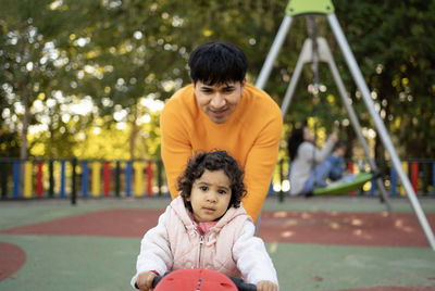 Young adult father pushing baby daughter on toy moto in playground