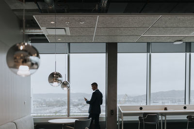 Rear view of businessman with smart phone standing by window at office