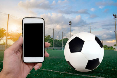 Close-up of hand holding smart phone against soccer ball on field