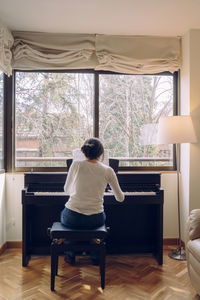 Rear view of woman playing piano while sitting on chair at home