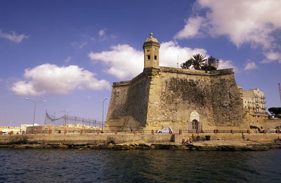 Low angle view of fort in front of river against cloudy sky on sunny day