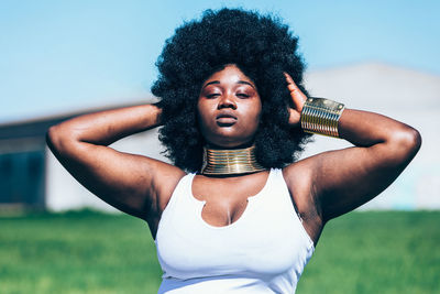 Curvy black female in trendy white dress with traditional african accessories and curly hair adjusting bracelet while standing on grass on cloudless day in field