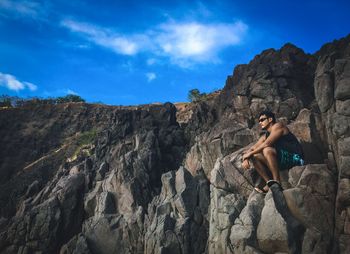 Low angle view of man sitting on rock formation against blue sky