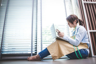 Side view of man using mobile phone while sitting on window