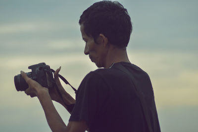 Side view portrait of young man holding camera against sky