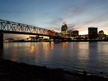 Illuminated bridge over river by buildings against sky at night