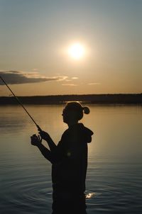 Side view of woman fishing in lake against sky during sunset