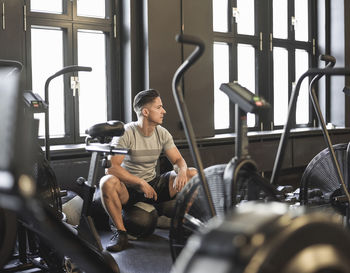 Full length of man looking away while sitting on equipment in gym