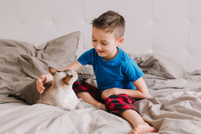 Caucasian boy sitting on bed in bedroom at home and petting stroking oriental cat. 