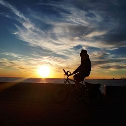 Silhouette man riding bicycle on beach against sky during sunset