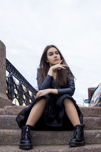 Young woman looking away while sitting on steps against sky