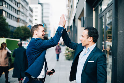 Happy mature male business coworkers giving high-five while standing on sidewalk in city