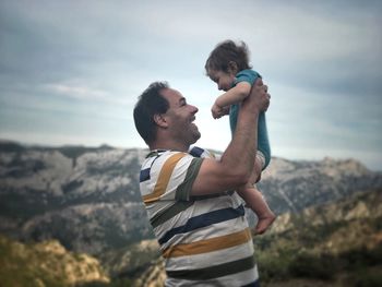 Side view of happy man carrying baby while standing against mountains