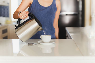 Woman pouring hot water from kettle in cup indoors.
