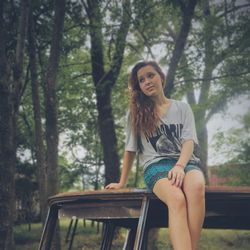 Beautiful young woman sitting on abandoned vehicle in forest