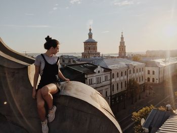 Woman sitting on building in city against sky