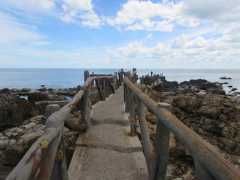 Footbridge on rocky coastline by sea against cloudy sky