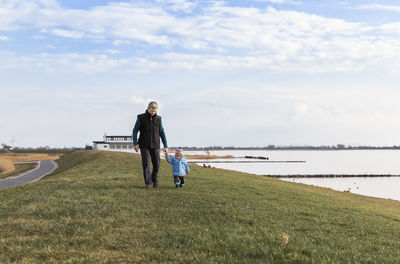 Grandfather and granddaughter walking by lake against sky