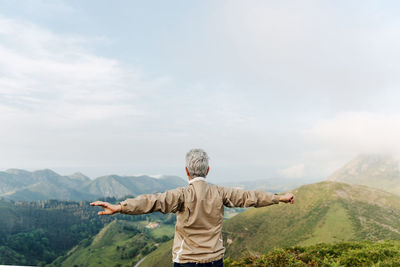 Back view of unrecognizable senior female traveler raising and stretching arms in morning in hilly terrain