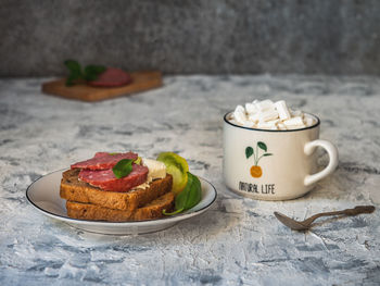 Smoked sausage and cheese sandwiches on toasted bread, tomato wedges and coffee in a white mug